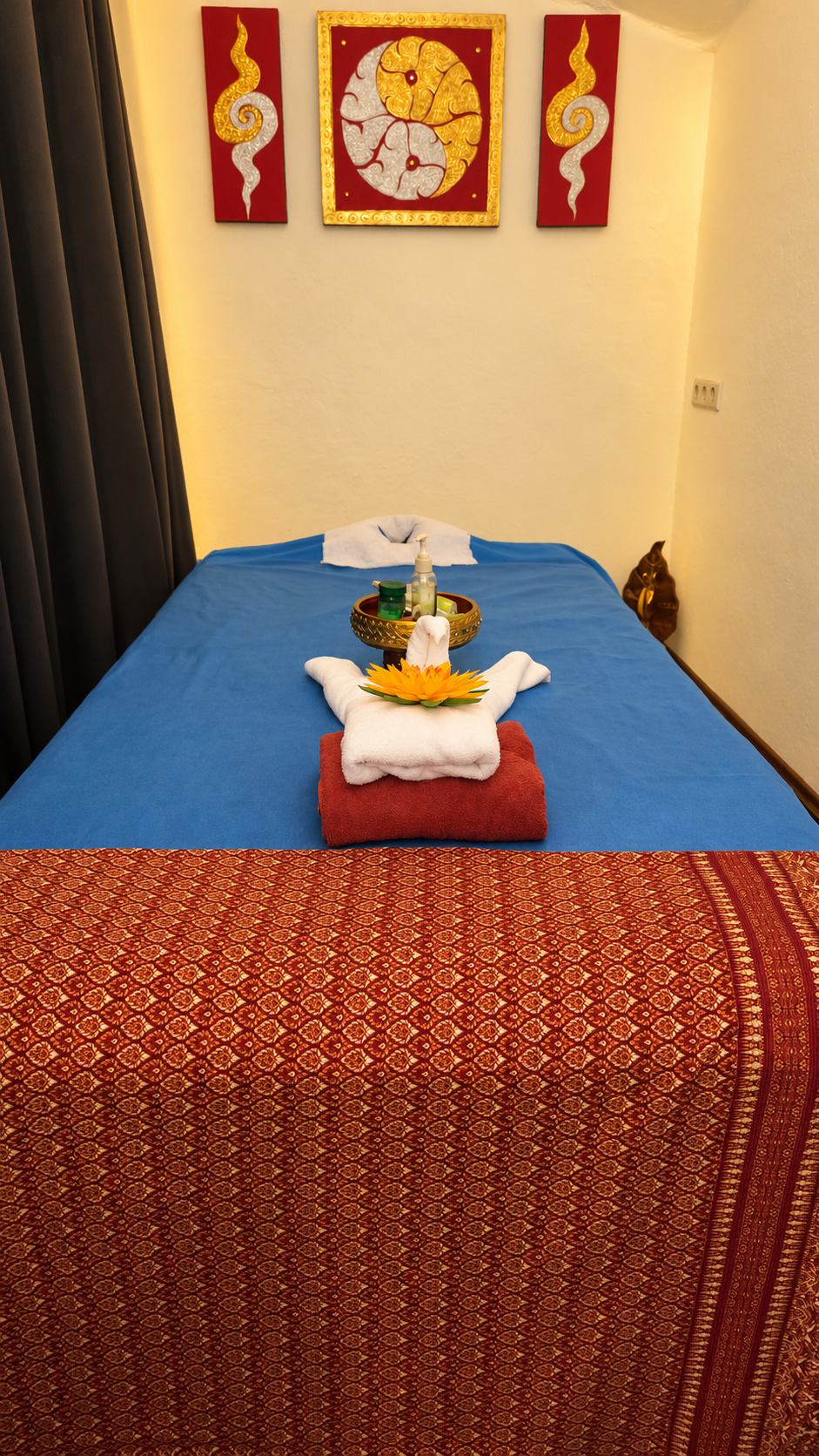 Massagebereich mit blauer Liege, thailändischen Textilien und gold-roter Wanddekoration